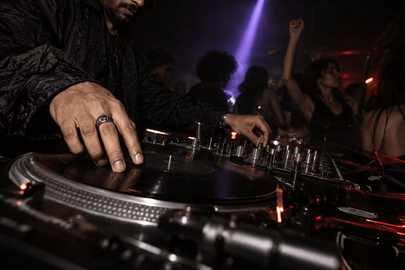 Hands on a turntable with dancers beyond the DJ booth